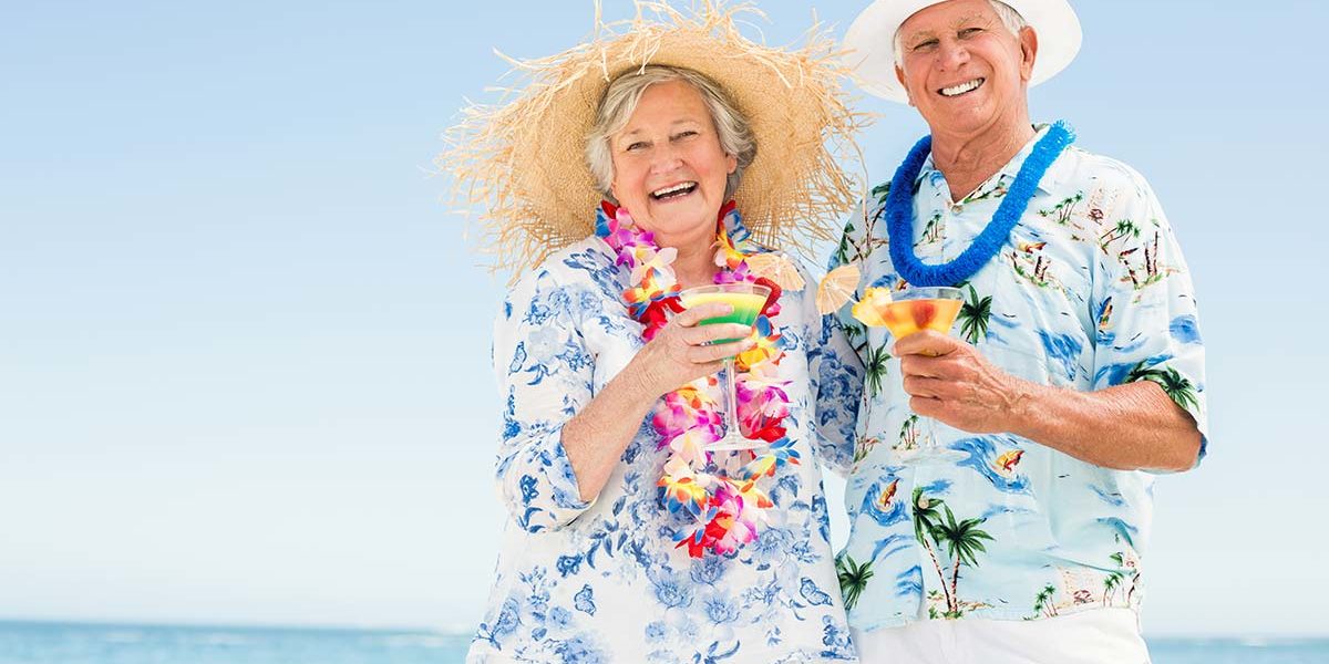Seniorenehepaar mit Blumenketten, Hawaiihemden und Sonnenhüten genießt Cocktails am Strand | Wohnen im Ausland