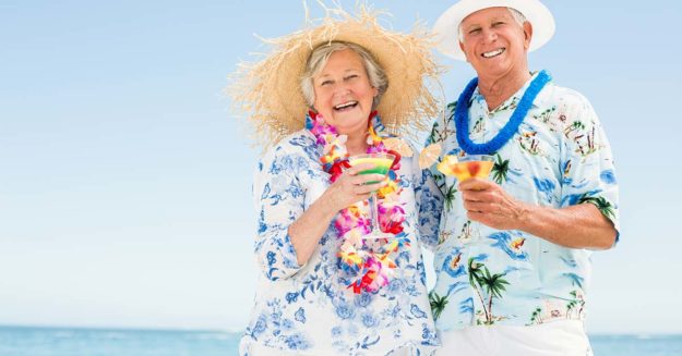 Seniorenehepaar mit Blumenketten, Hawaiihemden und Sonnenhüten genießt Cocktails am Strand | Wohnen im Ausland