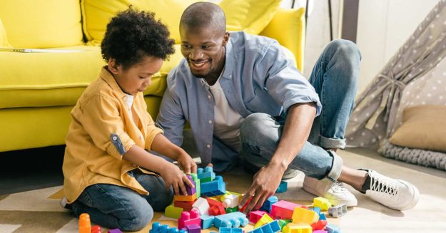 Vater und Sohn sitzen vor gelbem Sofa auf dem Boden und spielen mit Bauklötzen - Scheidungsimmobilie