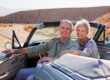 Ein Seniorenpaar sitzt in einem Cabrio auf einer Landstraße in einer weiten Landschaft, die Fahrertür ist auf und beide blicken nach hinten | Immobilienverrentung