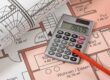 Auf dem Grundrissplan einer Immobilie liegen ein weiterer zusammengerollter Grundrissplan, ein Taschenrechner und ein Stift | Immobilienfinanzierung