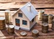 Modell eines Hauses steht auf einem Holztisch und darum stehen kleine Türmchen aus Ein-Euro-Münzen | Immobilienfinanzierung