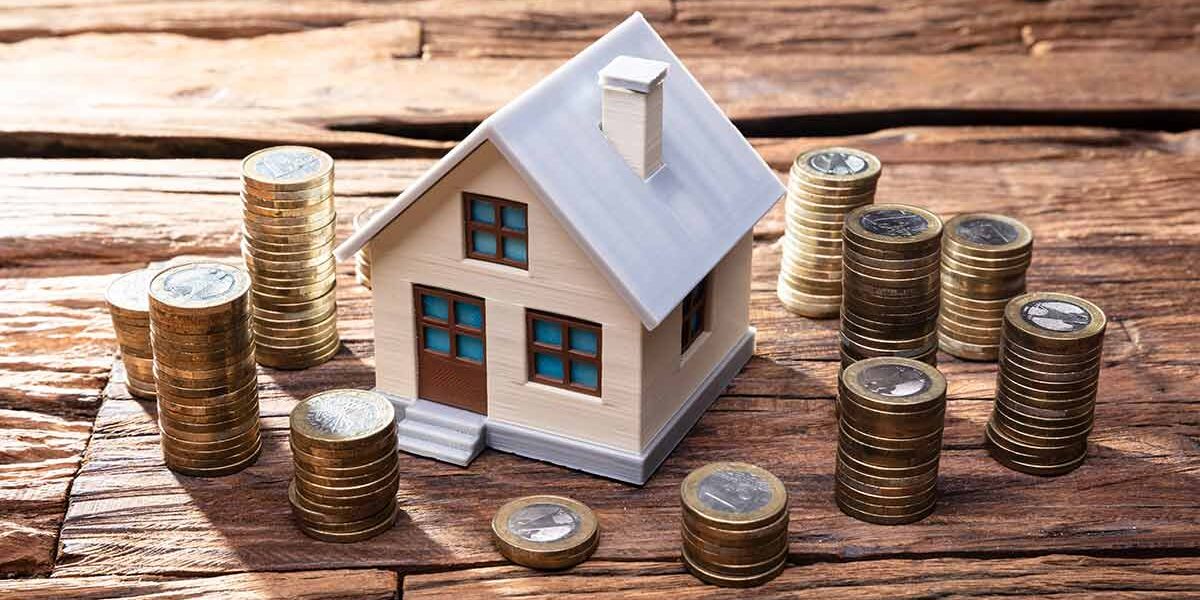 Modell eines Hauses steht auf einem Holztisch und darum stehen kleine Türmchen aus Ein-Euro-Münzen | Immobilienfinanzierung