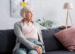 Eine Seniorin sitzt auf ihrem Sofa in ihrem Wohnzimmer, im Hintergrund hängt ein modernes Gemälde, daneben stehen eine Topfpflanze, eine Stehlampe und ein Regal | Wohnen im Alter